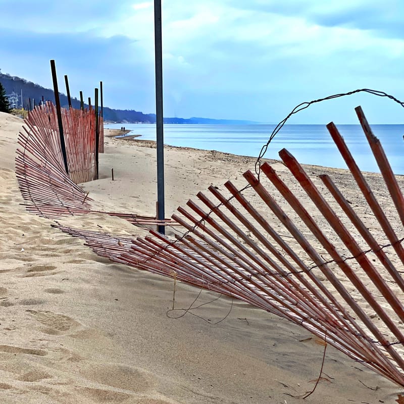 A weathered wood dune fence curves along a sandy beach beneath a pale blue sky