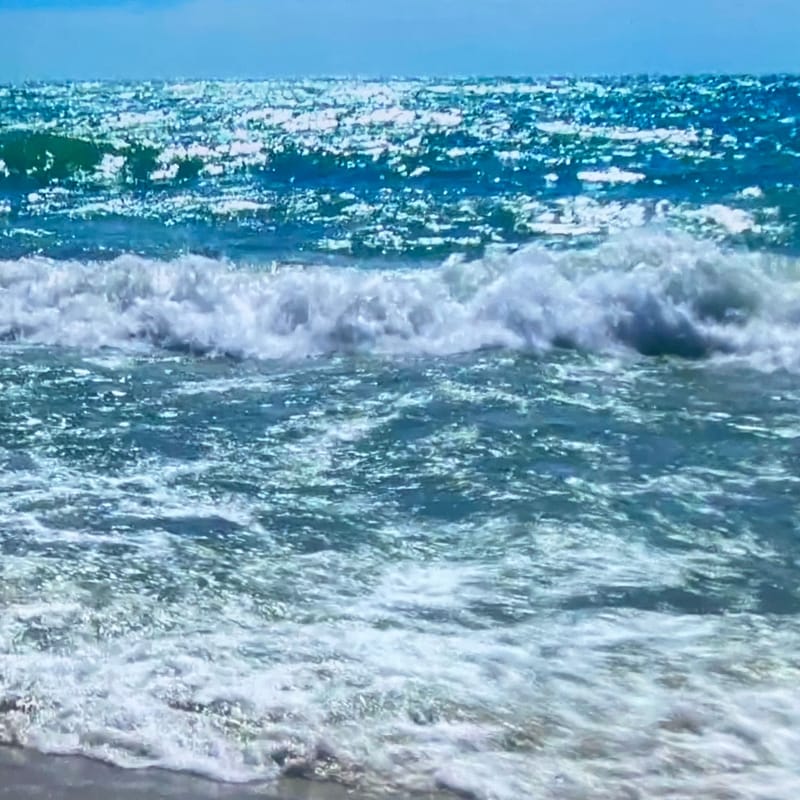 Foamy white wave crashing onto gray sand with teal ocean beyond
