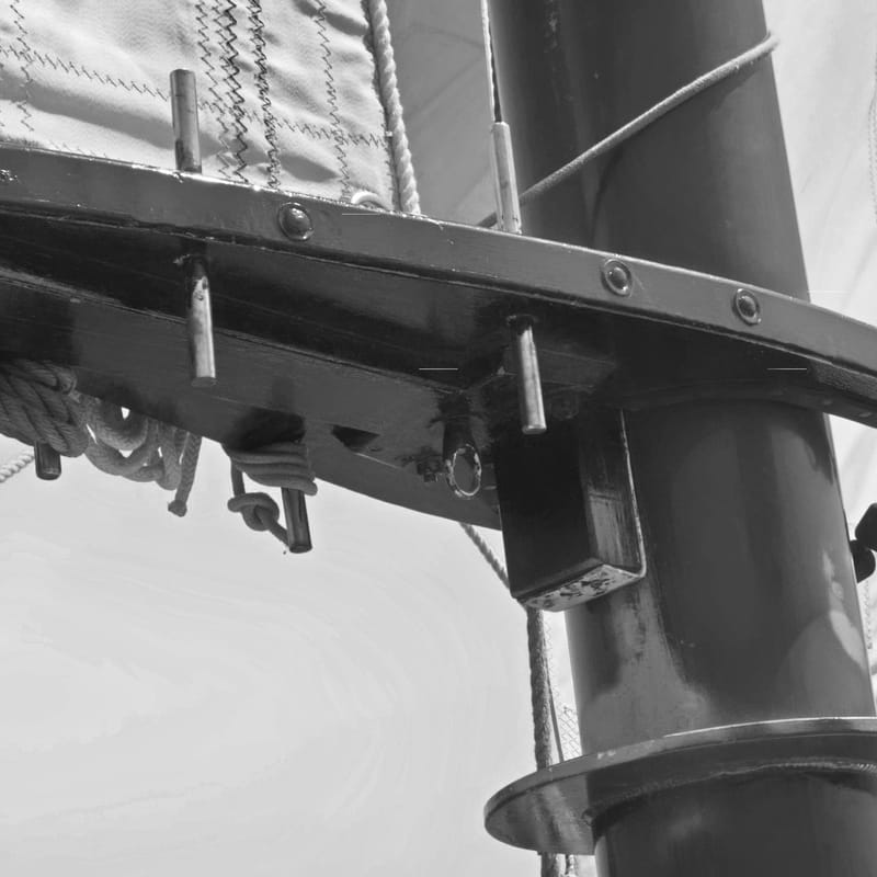 Black-and-white close-up of a boom fitting and belaying pins where mast meets spar, sail behind