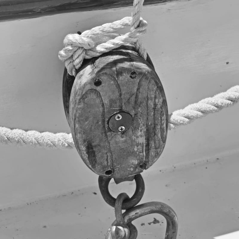 Black-and-white close-up of a weathered wooden ship's pulley wrapped in rope, iron chain and shackle hanging below