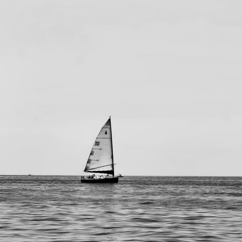 Black-and-white minimalist seascape of a small sailboat under full sail on open water beneath a pale sky