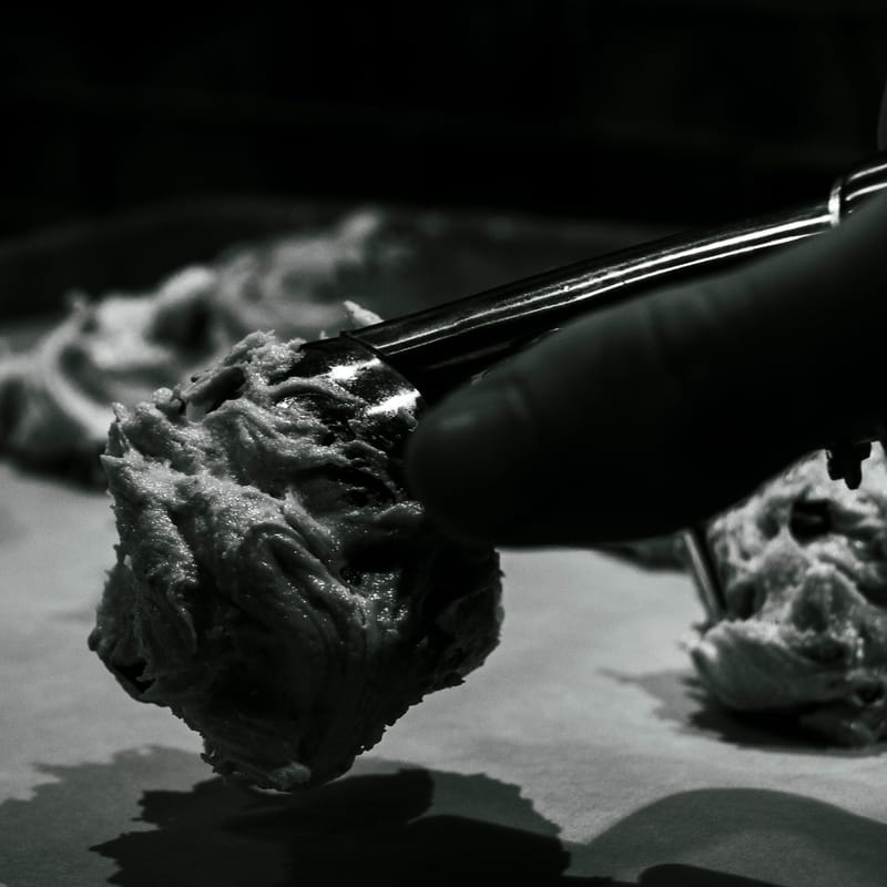 Black-and-white close-up of a gloved hand scooping cookie dough onto a baking sheet, dramatic side lighting